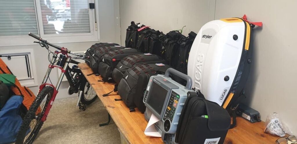 Matériel de secours professionnel composé d’un scope Corpuls 3 pour la surveillance cardiaque, de sacs à dos Dimatex rouges pour le transport du matériel médical, et d’une planche à masser utilisée lors des interventions d’urgence.